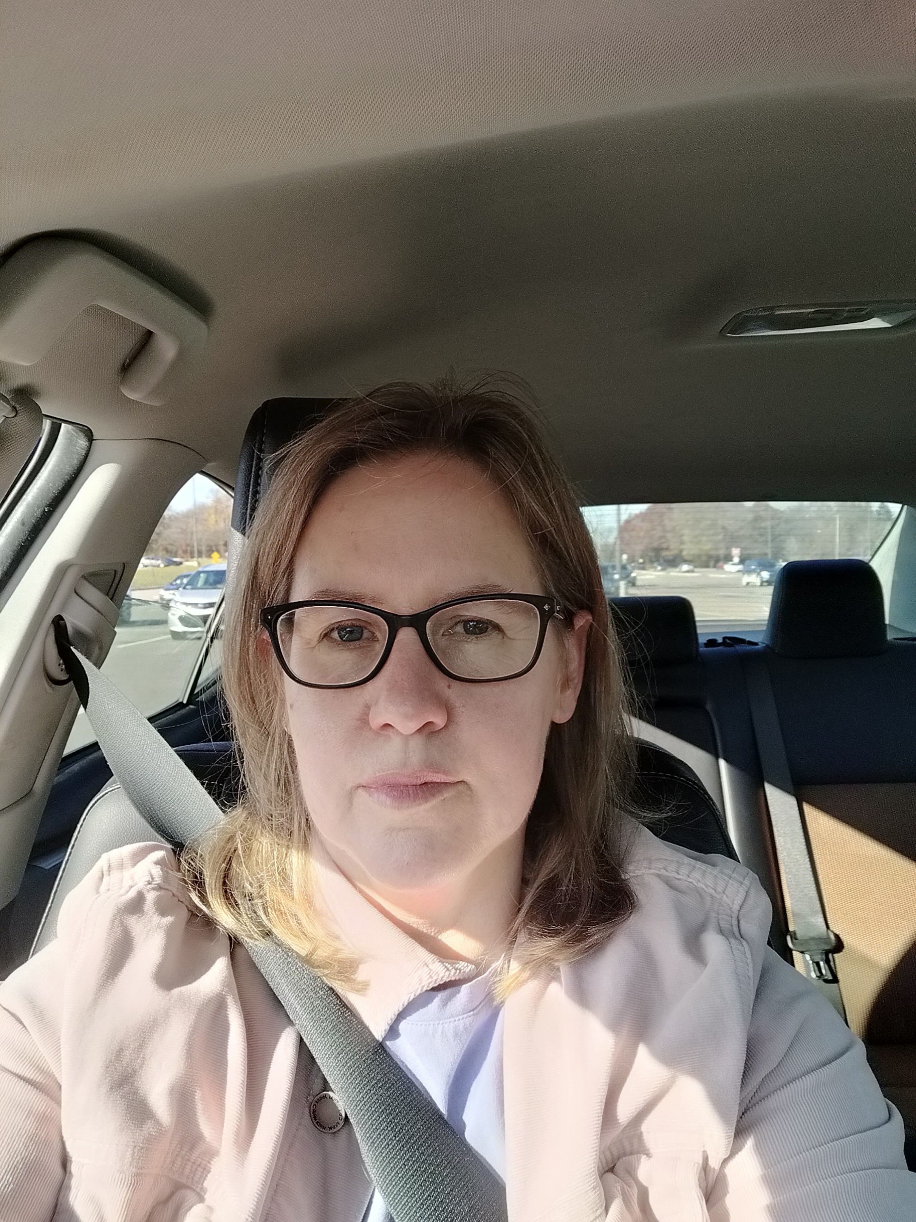 Woman with light brown hair and dark rimmed glasses sitting in car with seat belt and neutral facial expression. 